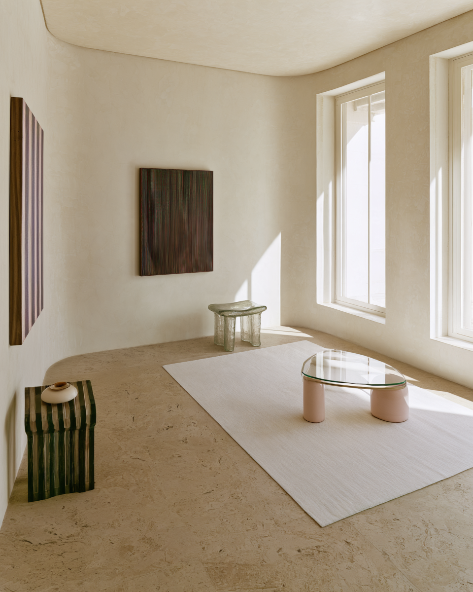 A minimalist interior room with textured beige plaster walls. Strata, a textured abstract painting with fine vertical raised lines in dark green, burgundy, bronze and earthy tones, framed in dark wood.